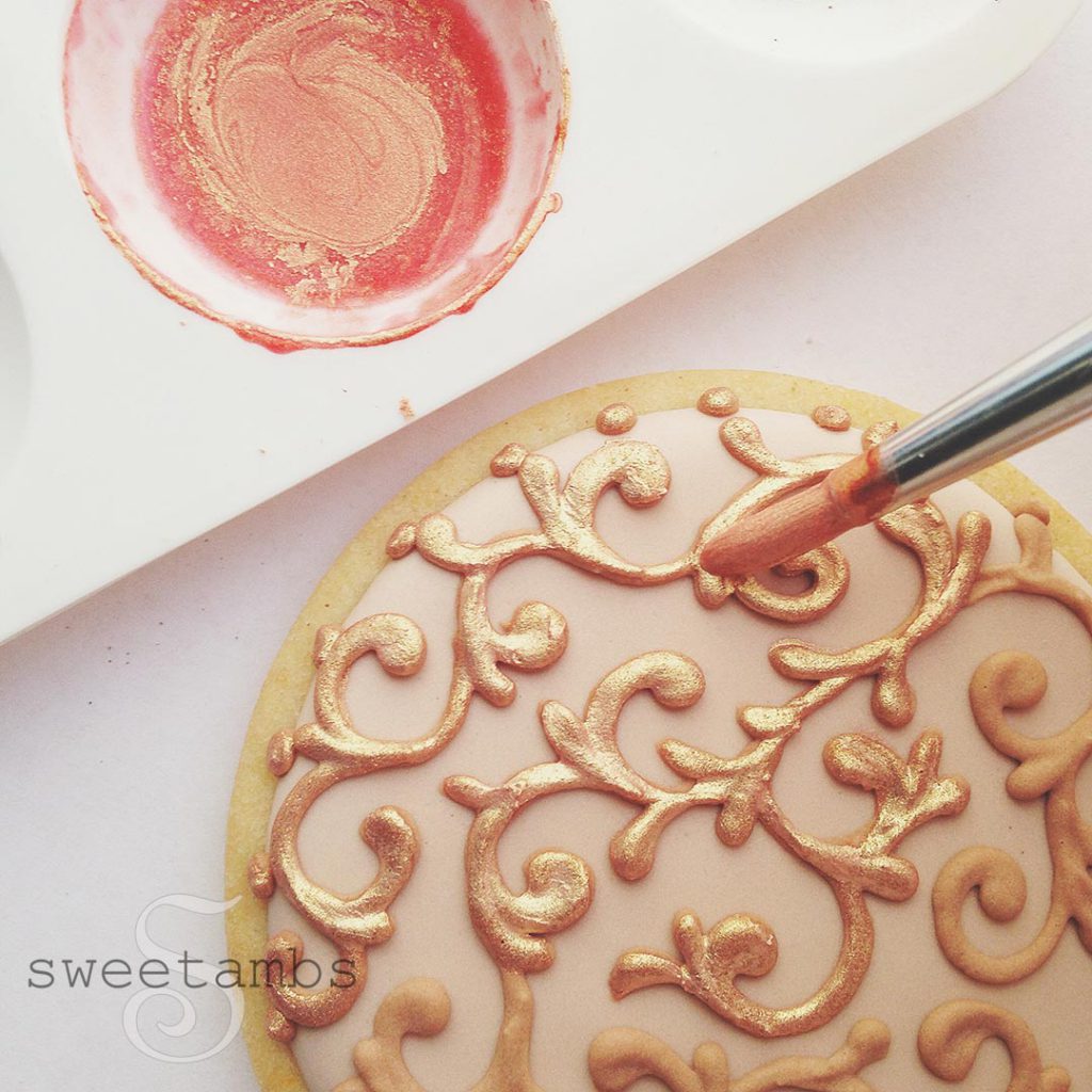 A brush dipped in liquid gold paint applying the gold to a filigree iced cookie.