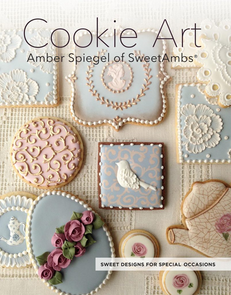 cookie decorating book cover by Amber Spiegel of SweetAmbs shows a flat lay of intricately decorated cookies in ivory, white, muted pink, blue, and green. The cookies are on a lace fabric background. The text on the cover reads Cookie Art Amber Spiegel of SweetAmbs Sweet Designs for Special Occasions 