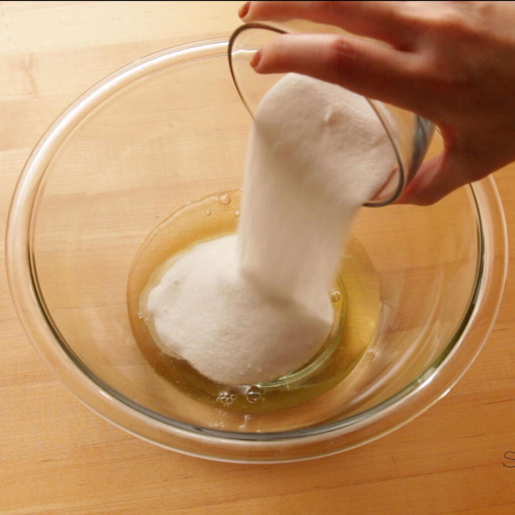 A hand is holding a small glass bowl pouring sugar into a larger glass bowl that has egg whites in it. The bowl is on a butcher block.