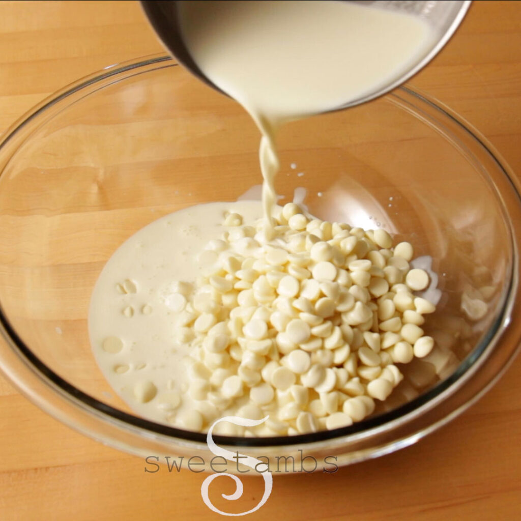 Heated heavy cream is being poured into a glass bowl full of white chocolate chips. The bowl is on a butcher block table.