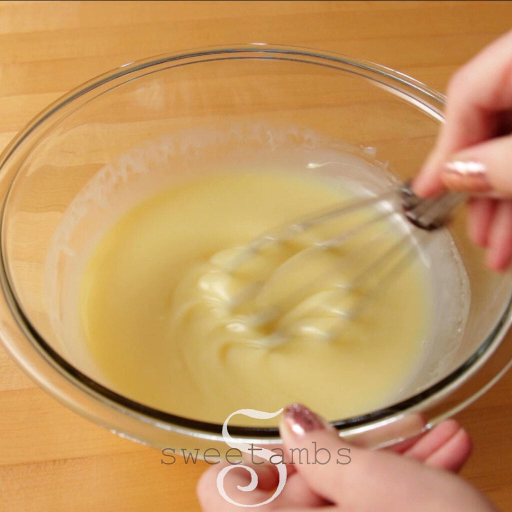 A glass bowl of liquid ganache is being whisked by hand while the other hand holds it steady. The bowl is on a butcher block table.