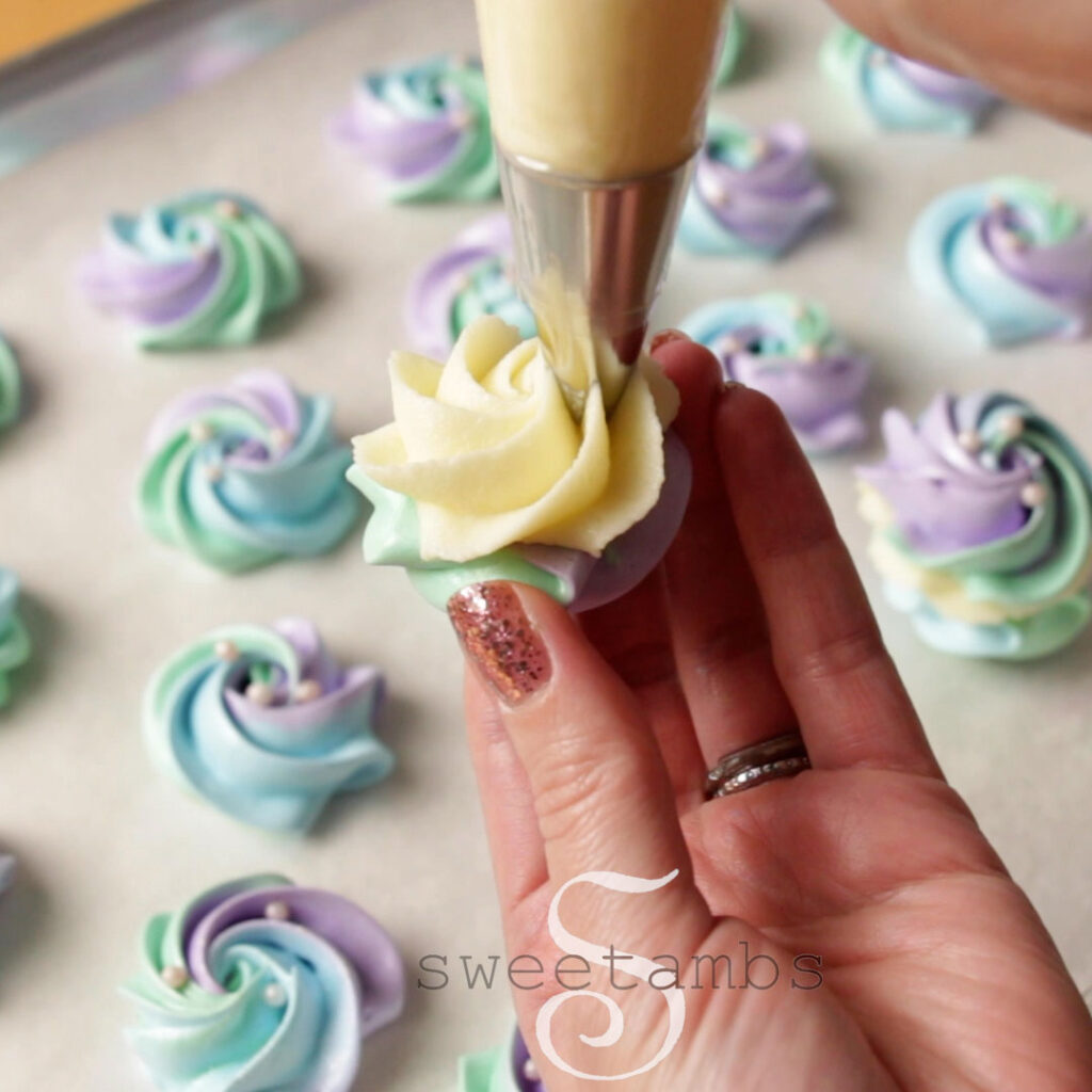 A hand is holding a meringue cookie while a swirl of white chocolate ganache is being piped on top. There is a tray of more meringue cookies in the background.