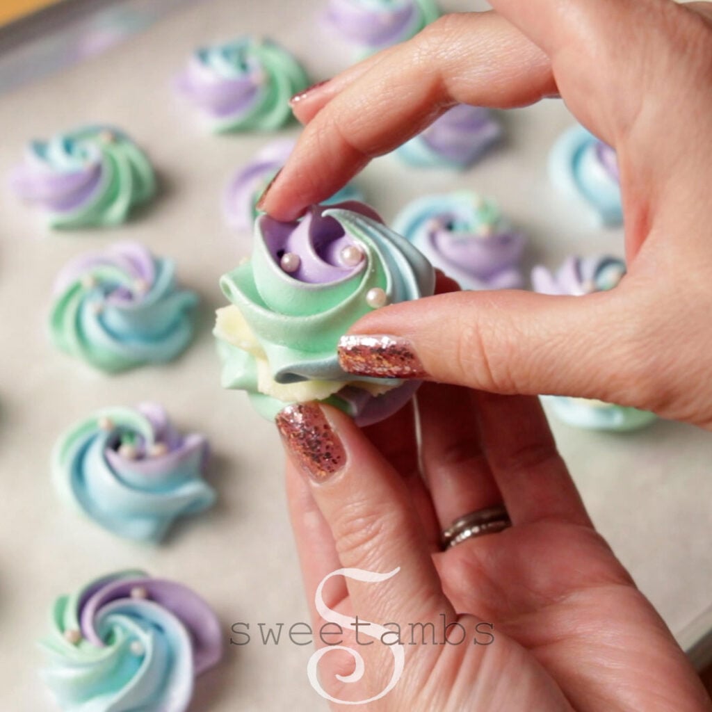 Two hands sandwiching two meringue cookies together white white chocolate ganache in between.