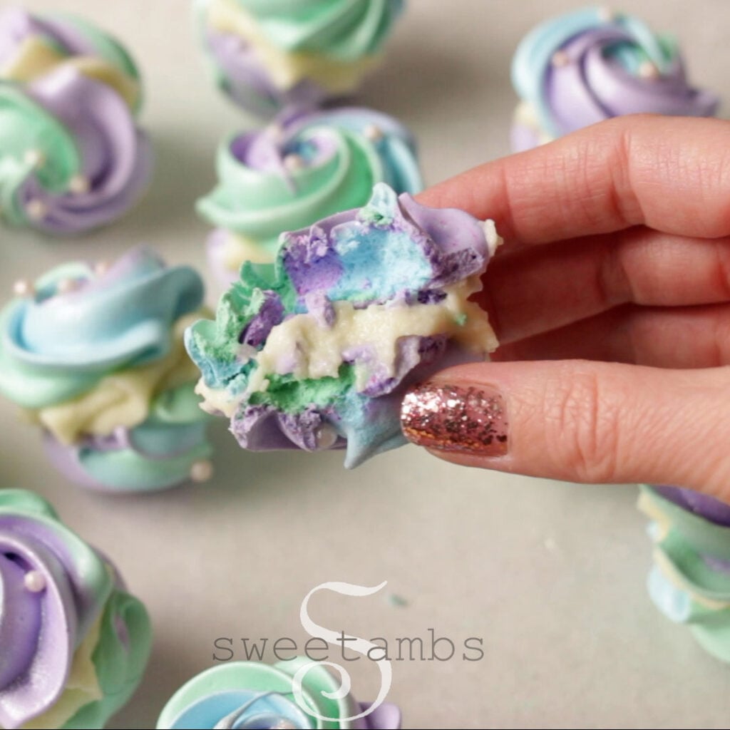 A hand is holding a meringue cookie sandwich with a bite taken out of it. There are more meringue cookie sandwiches in the background.