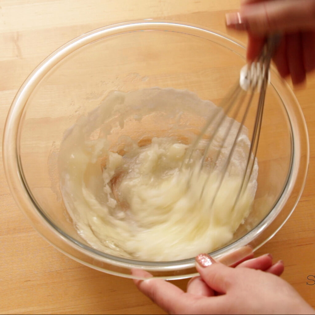 A hand is holding a glass bowl filled with egg whites and sugar while the other hand whisks it together. The bowl is on a butcher block table.