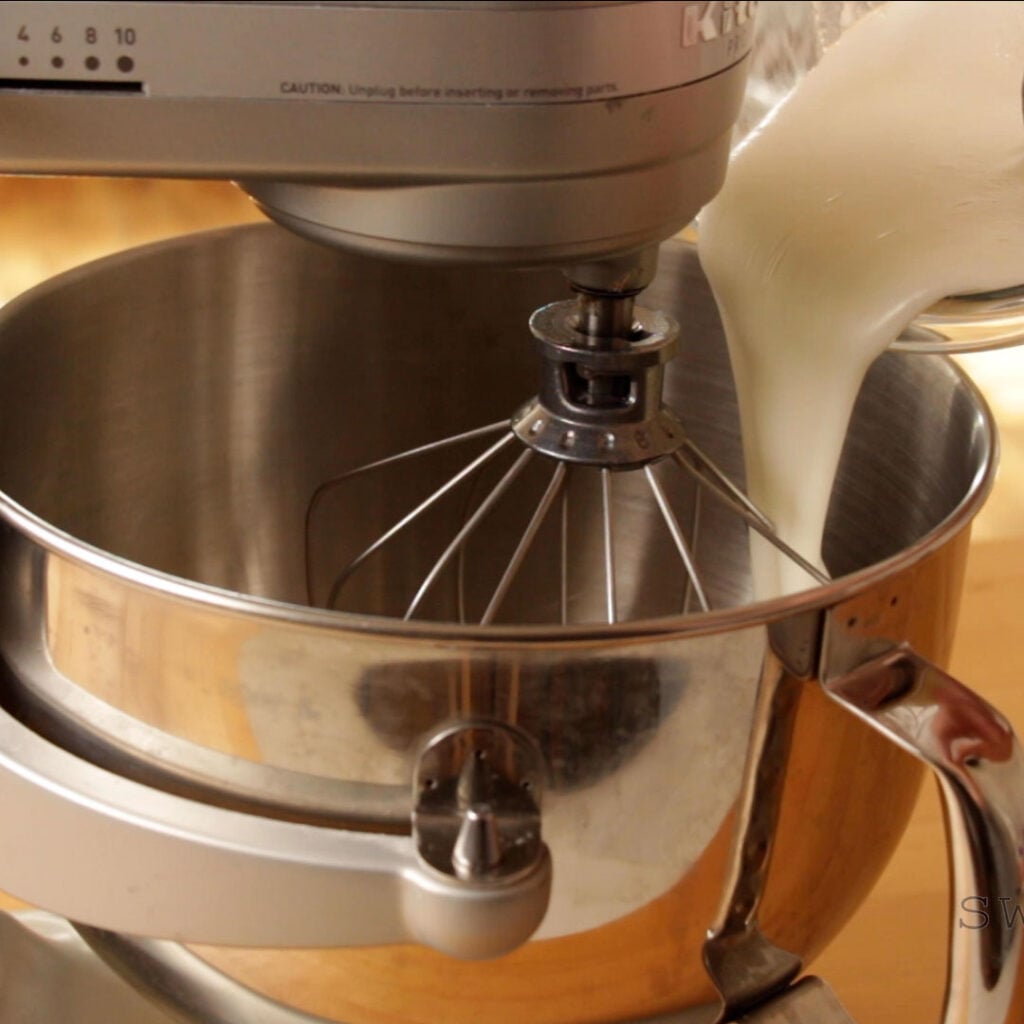 Foamy egg whites and sugar are being poured into the bowl of a stand mixer fitted with a whisk attachment.