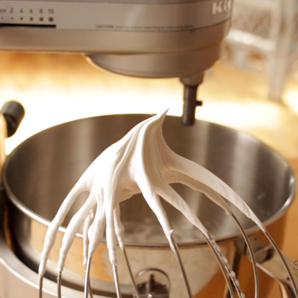 A whisk attachment from a stand mixer has a dollop of stiff peak meringue on it. There is a stand mixer in the background.