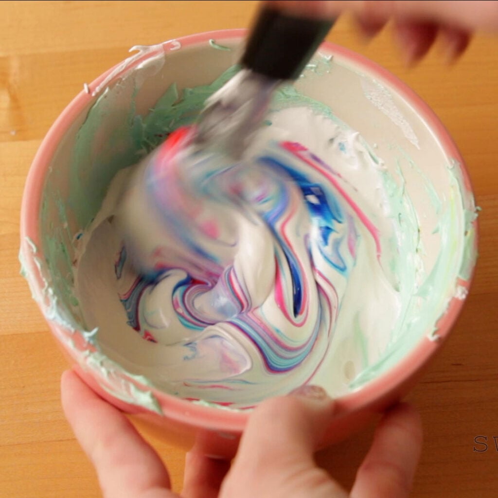 A bowl of meringue with swirls of pink and blue food coloring. One hand is holding the bowl steady while the other hand mixes in the color with a metal spatula.