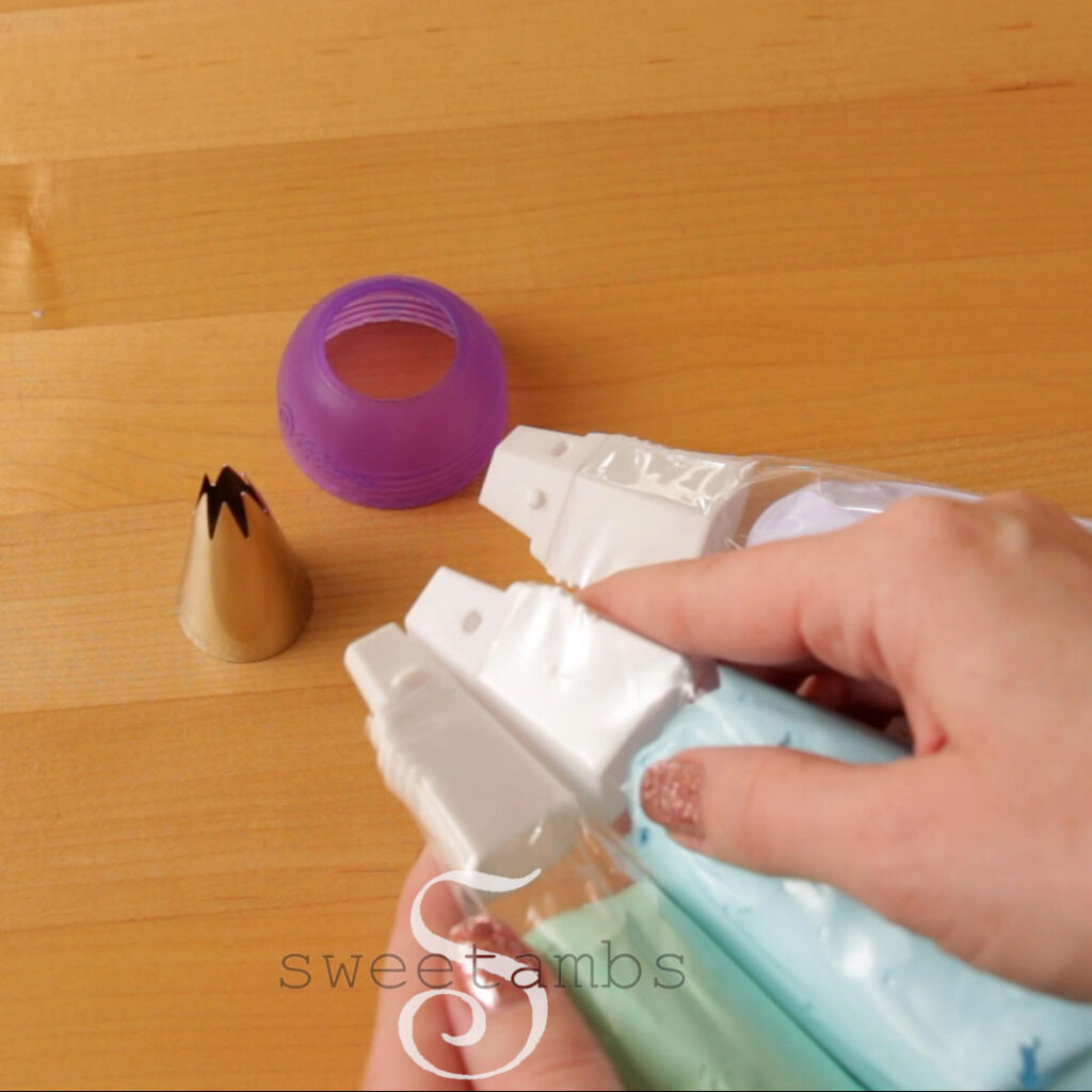3 coupler pieces of a 3-color coupler are inside of decorating bags, each filled with a different color of meringue - pastel green, blue, and purple. A star tip 1M and the cap from the 3-color coupler are on the table next to the decorating bags. 