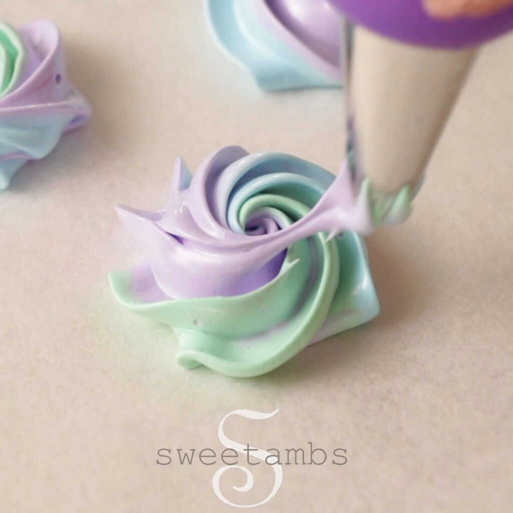 a 1M star tip is piping a swirl of meringue on a parchment lined baking sheet. The meringue is pastel purple, green, and blue.