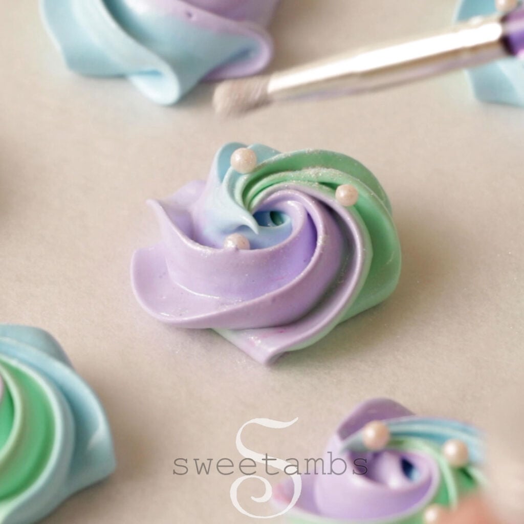 a brush is sprinkling white pearl dust on to a meringue cookie. There are 3 white sugar pearls on the meringue cookie.