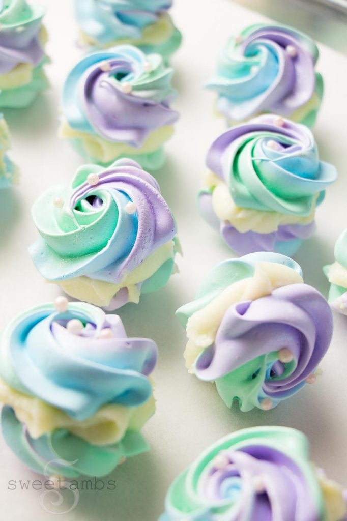 a ¾ view of meringue cookies on a parchment lined baking sheet. The meringue cookies are swirled with pastel purple, blue, and green and decorated with edible pearls and luster dust. They are sandwiched with white chocolate ganache.