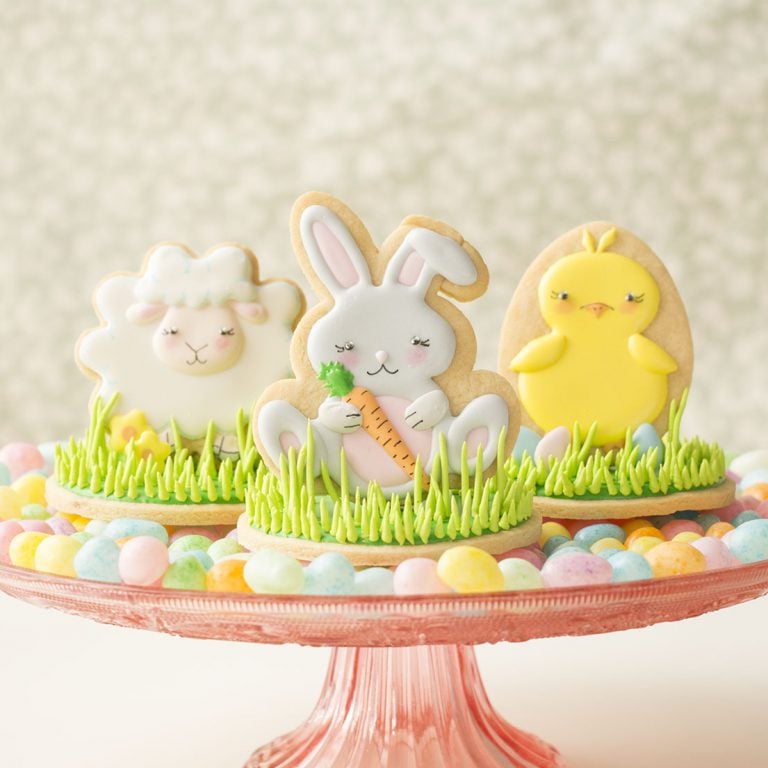 Easter cookies decorated to look like a lamb, baby bunny, and a chick. The cookies are standing upright on a cake platter surrounded by candy eggs