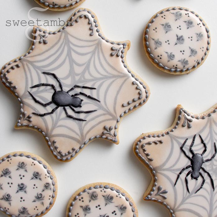 Plaque shaped Halloween cookies decorated in beige icing with inlaid gray spider webs. There are black royal icing spiders on the cookies and delicate borders on the edges. There are small round cookies decorated with beige icing and gray roses.
