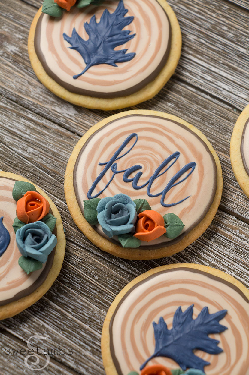 Fall cookies decorated to look like sliced branches. The cookie in the center says fall in cursive. There are two cookies decorated with navy blue oak leaves. The cookies are decorated with orange and blue royal icing roses and green royal icing leaves.