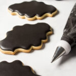 Plaque shaped cookies decorated with black royal icing. There is a decorating bag with a coupler and tip filled with black icing next to the cookies.