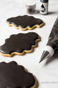 Plaque shaped cookies decorated with black royal icing. There is a decorating bag fitted with a coupler and a metal decorating tip filled with black icing lying next to the cookies. In the background is a bottle of black food coloring and a jar of black powdered food coloring.