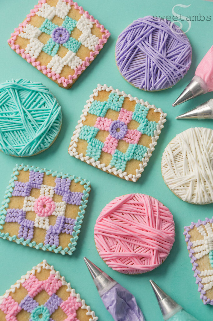 A set of cookies decorated to look like crochet granny squares and balls of yarn. There are decorating bags with metal tips filled with icing on the edges of the picture.
