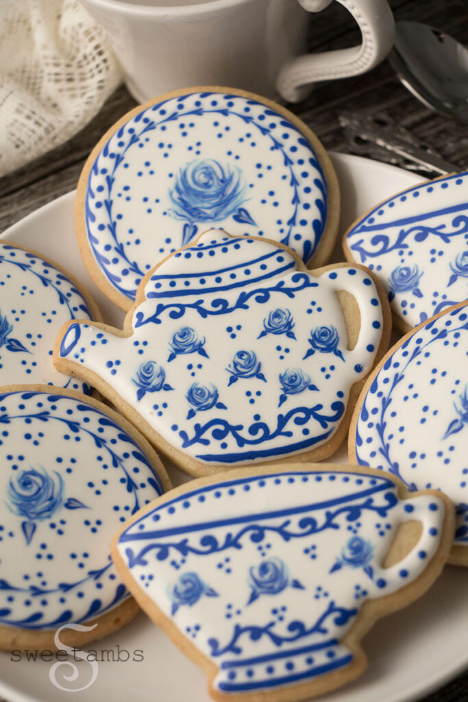 cookies decorated to look like a Delft pottery tea set. The cookies are on a white plate on a wood surface. There are silver teaspoons and the handle of a teacup next to the plate. There is an antique style lace fabric behind the cookies.