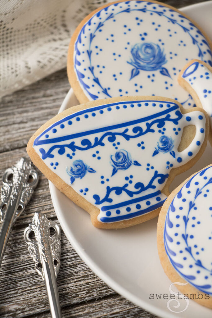 cookies decorated to look like a Delft pottery tea set. The cookies are on a white plate on a wood surface. There are silver teaspoons and the handle of a teacup next to the plate. There is an antique style lace fabric behind the cookies.