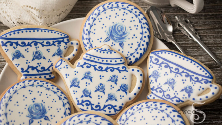 cookies decorated to look like a Delft pottery tea set. The cookies are on a white plate on a wood surface. There are silver teaspoons and the handle of a teacup next to the plate. There is an antique style lace fabric behind the cookies.