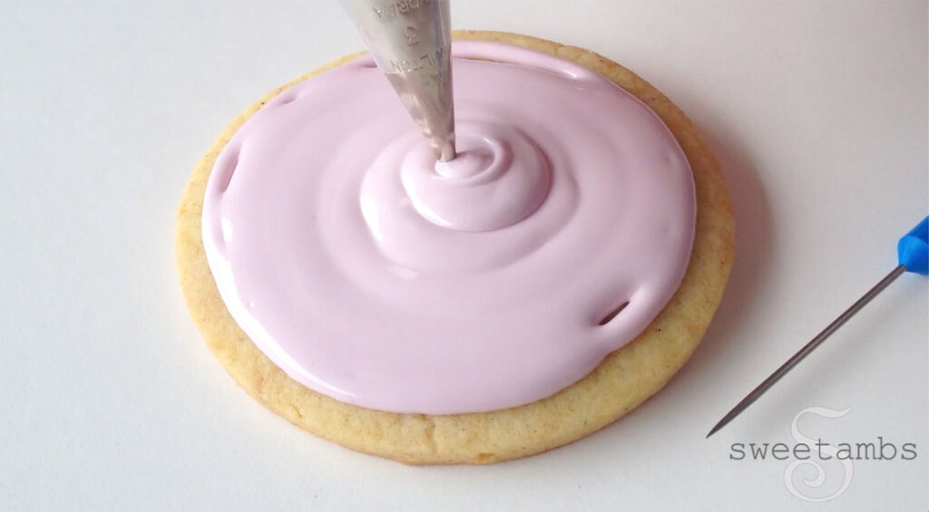 Flooding a cookie with royal icing. A decorating tip in the center of the cookie is applying flood consistency royal icing. There is a scribe tool, which is a needle with a blue plastic handle, next to the cookie.