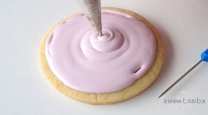 A round cookie being iced with light pink flood consistency royal icing. There is a metal decorating tip in the middle of the cookie applying the icing. There is a scribe tool (needle with blue handle) next to the cookie.