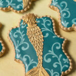 A plaque shaped cookie decorated with a filigree design and a gold mermaid. There is a gold bead border around the edge of the cookie.