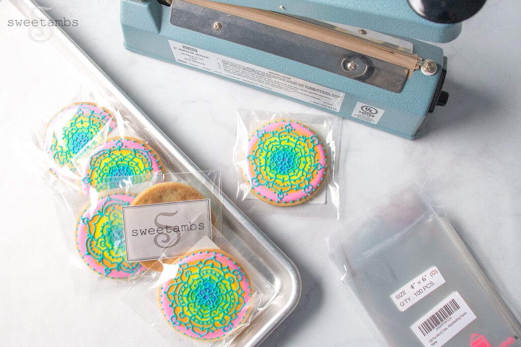 Brightly colored decorated cookies wrapped in cello bags on a baking sheet. One cookie is off of the baking sheet sitting in front of a heat sealer. There are empty cello bags next to the tray of cookies.