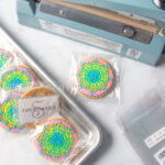 Brightly colored decorated cookies wrapped in cello bags on a baking sheet. One cookie is off of the baking sheet sitting in front of a heat sealer. There are empty cello bags next to the tray of cookies.