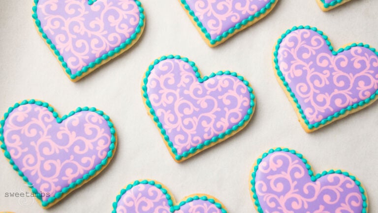 Heart shaped cookie decorated with pink and purple filigree and a teal bead border