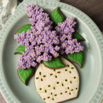 A set of cookies decorated to look like lilacs in an eyelet lace vase. The cookies are sitting on a platter on lace fabric and a wood background.