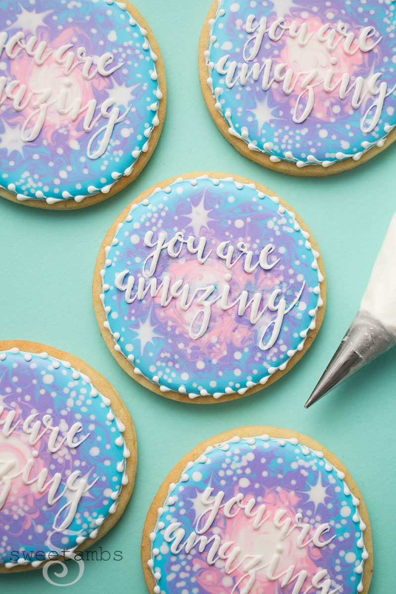 A piping bag with a decorating tip next to a set of galaxy cookies with the words "you are amazing" piped in icing.