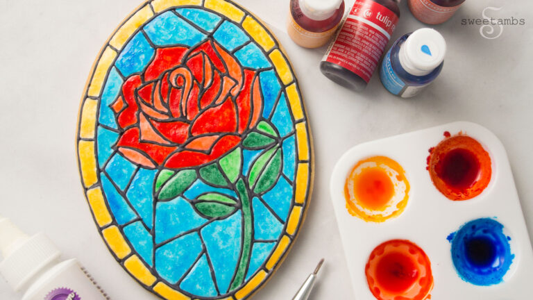 a cookie decorated to look like a stained glass rose. The cookie is surrounded by bottles of food coloring and a palette filled with food coloring.