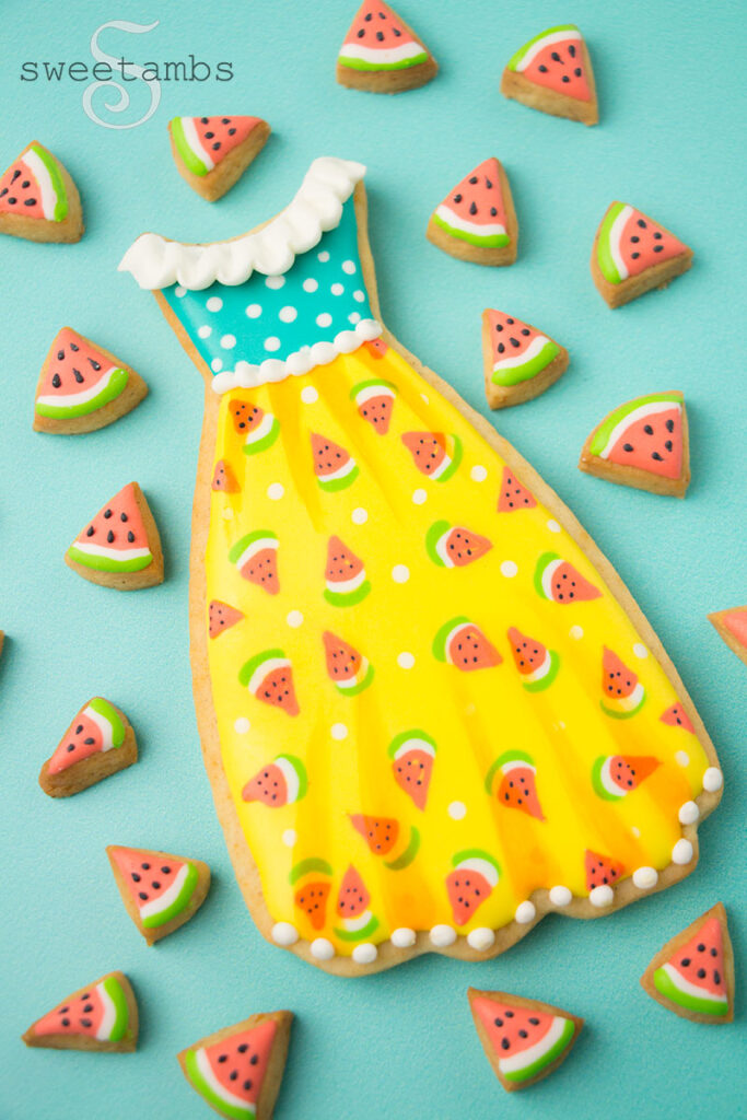 A sleeveless dress cookie decorated with a teal polka dot top and yellow skirt with little watermelons. There is a white ruffle along the neck of the dress. The dress is surrounded by tiny watermelon cookies.