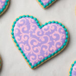 Heart shaped cookie decorated with pink and purple filigree and a teal bead border