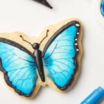 A butterfly cookie decorated with black royal icing and blue edible paint. The cookie is surrounded by a paint brush, an edible ink marker, a palette filled with blue edible paint, and a decorating bag filled with black icing.