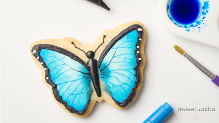 A butterfly cookie decorated with black royal icing and blue edible paint. The cookie is surrounded by a paint brush, an edible ink marker, a palette filled with blue edible paint, and a decorating bag filled with black icing.