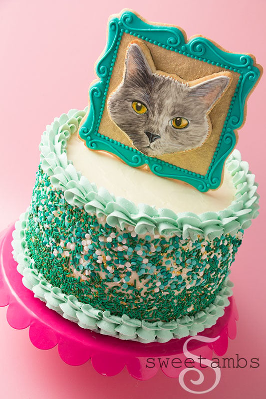 A cookie decorated to look like a portrait of a cake in a picture frame. The cookie frame is embellished with a gold background and filigree details around the edge. The cookie is sitting on top of a cake decorated with a shell border and sprinkles. The cake is on a scalloped cake stand.