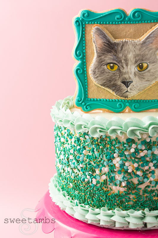 A cookie decorated to look like a portrait of a cake in a picture frame. The cookie frame is embellished with a gold background and filigree details around the edge. The cookie is sitting on top of a cake decorated with a shell border and sprinkles. The cake is on a scalloped cake stand.