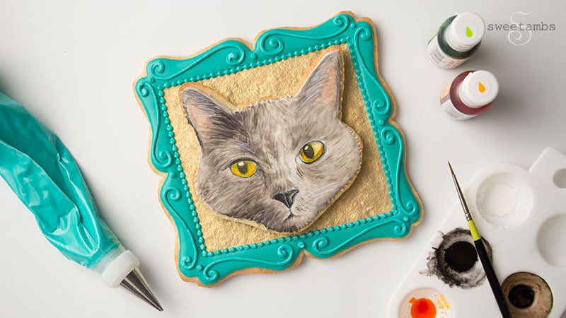 A cookie decorated to look like a portrait of a cake in a picture frame. The cookie frame is embellished with a gold background and filigree details around the edge. The cookie is surrounded by a decorating bag filled with icing, food coloring bottles, a paint palette, and a paint brush.