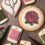 A set of cookies decorated to look like embroidery supplies including scissors, bobbins, and an embroidery hoop with a rose. There are tipless decorating bags next to the cookies filled with icing.