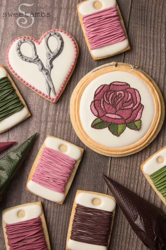 A set of cookies decorated to look like embroidery supplies including scissors, bobbins, and an embroidery hoop with a rose. There are tipless decorating bags next to the cookies filled with icing.