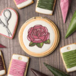 A set of cookies decorated to look like embroidery supplies including scissors, bobbins, and an embroidery hoop with a rose. There are tipless decorating bags next to the cookies filled with icing.