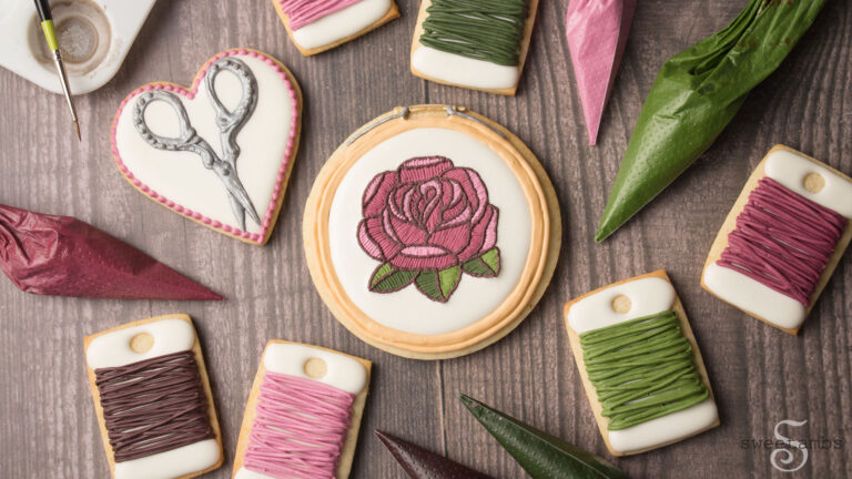 A set of cookies decorated to look like embroidery supplies including scissors, bobbins, and an embroidery hoop with a rose. There are tipless decorating bags next to the cookies filled with icing.
