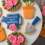 A set of cookies sitting on a plate with a wooden background. The cookies are decorated with a garden theme to look like flower pots, sprouts, dianthus, a watering can, a hand rake, and a shovel.