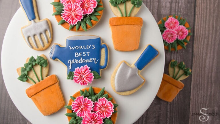 A set of cookies sitting on a plate with a wooden background. The cookies are decorated with a garden theme to look like flower pots, sprouts, dianthus, a watering can, a hand rake, and a shovel.