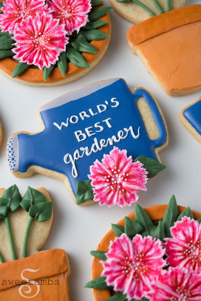 A cookie decorated to look like a watering can with a dianthus on it and the words "World's Best Gardener" written in icing. The cookie is surrounded by cookies decorated to look like terra cotta pots with sprouts and dianthus growing.