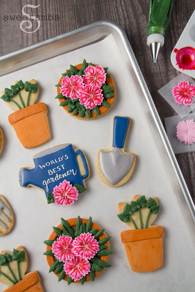 Cookies on a parchment-lined baking tray on a wooden background. The cookies are decorated in a garden theme to look like a watering can, flower pots, sprouts, dianthus, a hand rake, and a shovel. Next to the tray is a decorating bag with green icing, a paint palette with pink food coloring, and partially completed royal icing flowers on parchment paper squares.