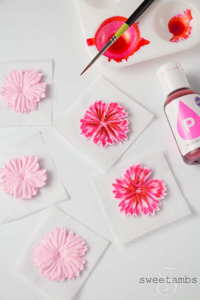 5 Royal icing flowers on parchment paper squares. 3 of the flowers are not painted and 2 of the flowers are painted with pink and red food coloring to add detail. There is a paint palette in the upper right corner filled with pink food coloring with a thin artist brush on the palette. There is a bottle of pink food coloring to the right of the flowers. 